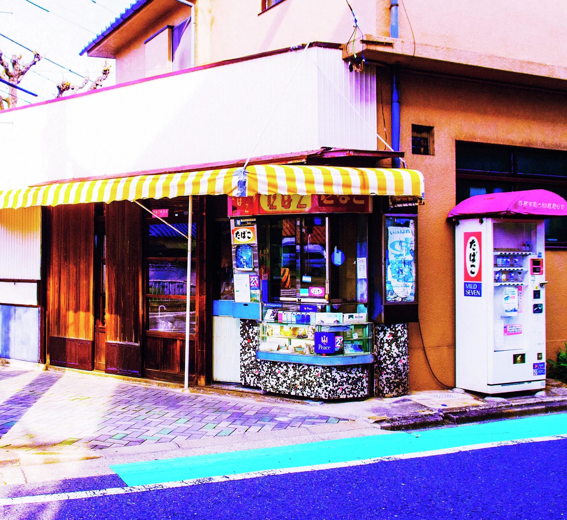 A striped yellow awning shades a corner shop beside a white vending machine with pink trim.