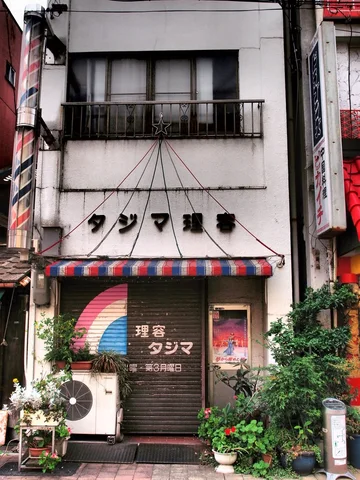 A narrow barber storefront has a striped awning, closed shutter, and lush potted plants below a balcony.