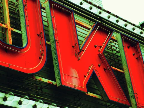 Oversized red metal letters project from green ribbed siding beneath a border of small bulbs.