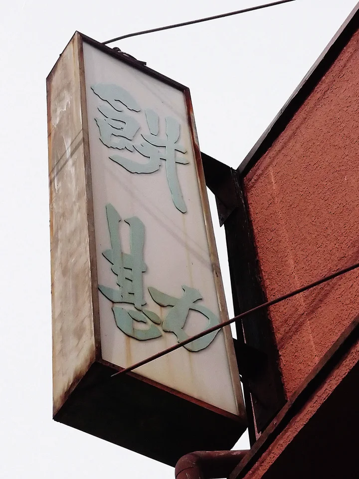 Faded raised Japanese characters cover a tall projecting rectangular sign beside a red brick building.