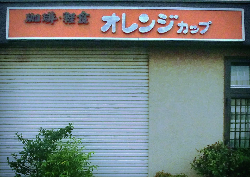 An orange Japanese sign hangs above a closed pale shutter beside shrubs and a barred window.