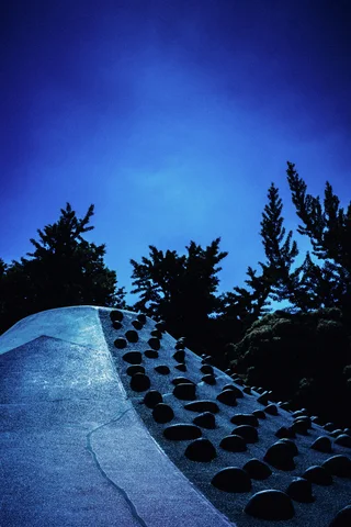 A blue sloping play structure with dark climbing holds rises before silhouetted trees against deep blue sky.