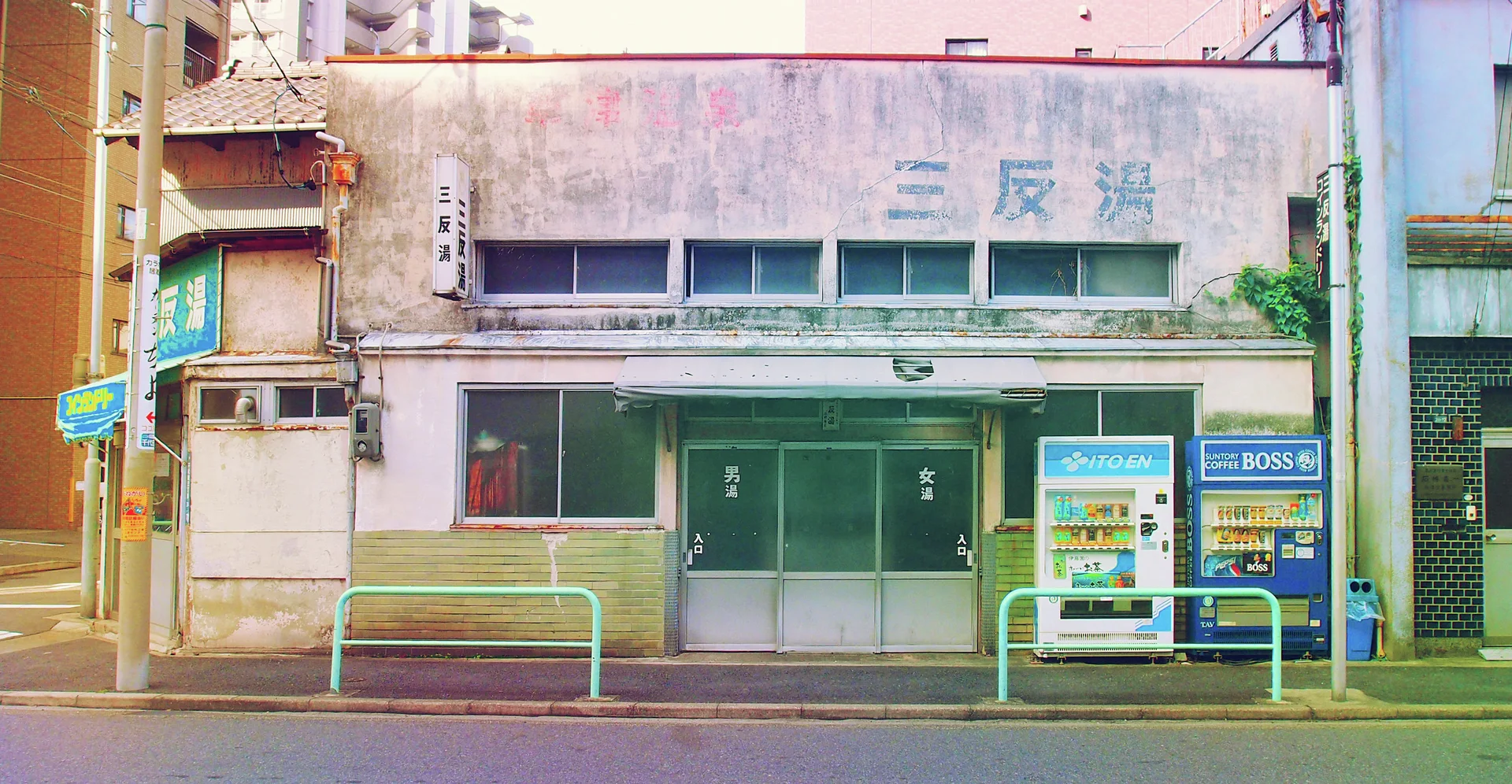 Two vending machines stand beside a low weathered building with green doors and pale turquoise roadside railings.