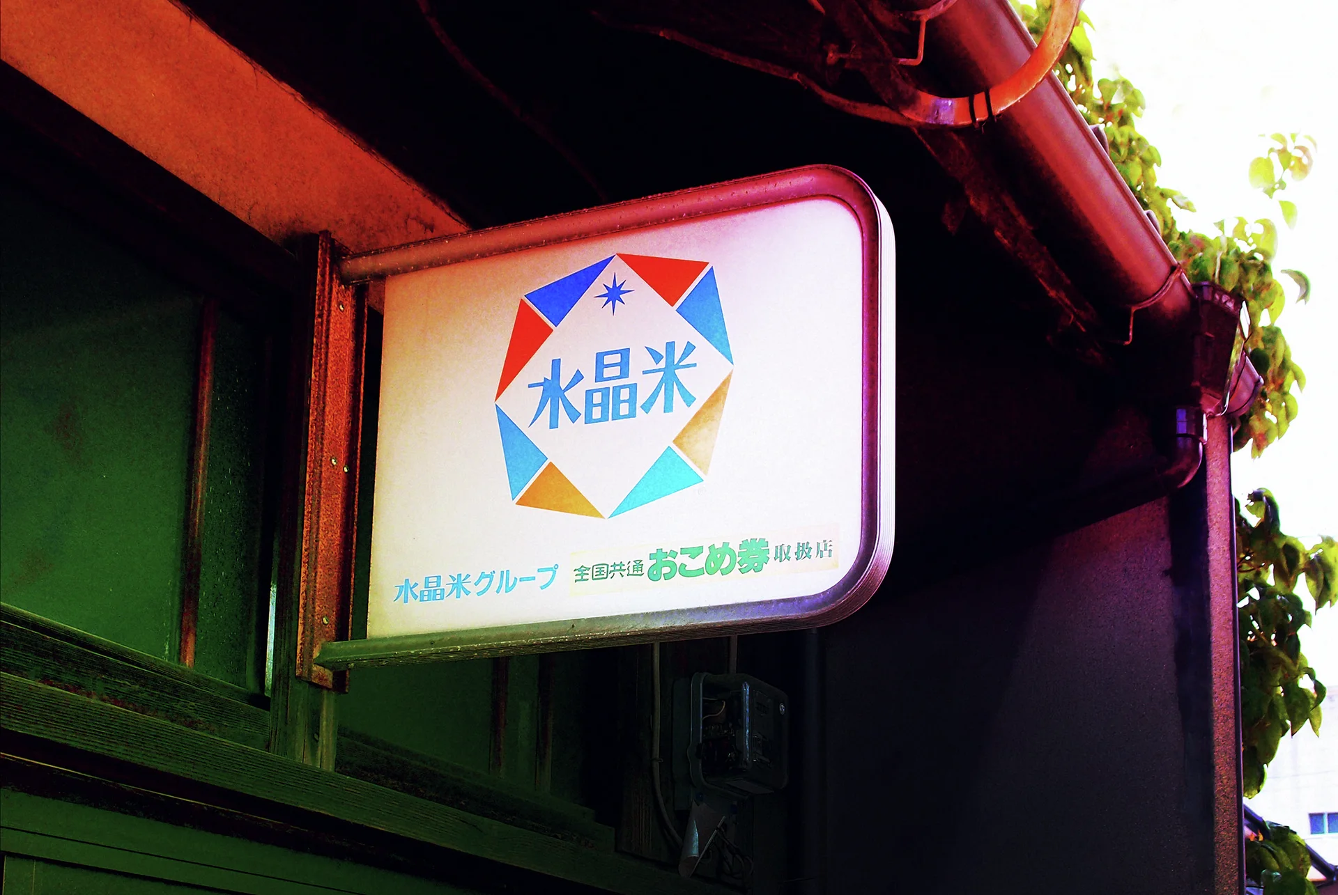 A square white hanging sign displays blue characters inside a multicolored geometric border beneath dark eaves.