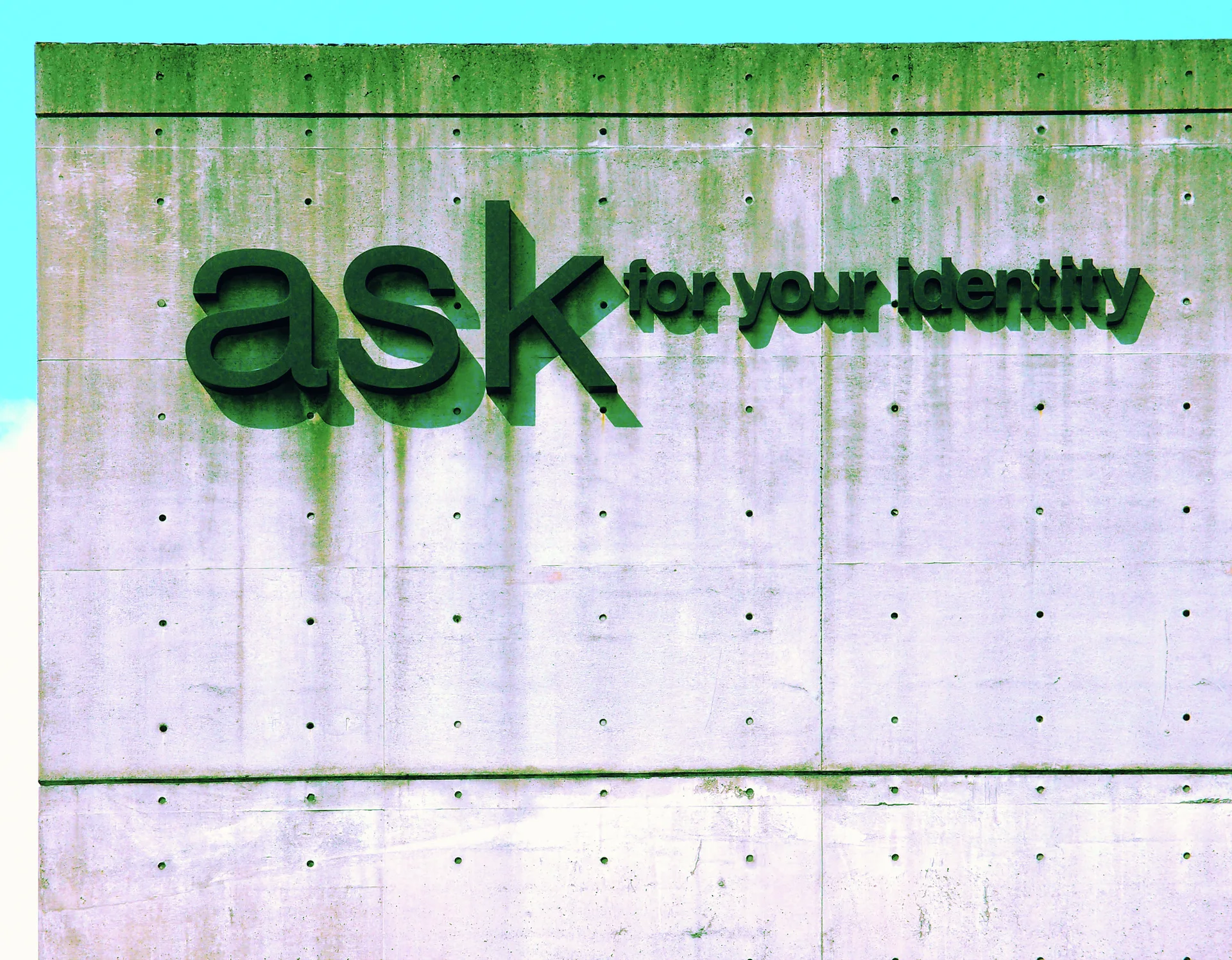 Raised green ask lettering casts shadows across a pale concrete wall with rows of small holes.