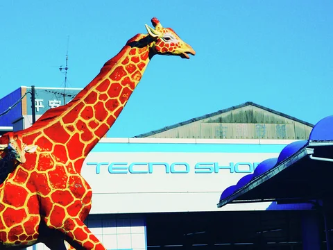 A bright orange giraffe sculpture rises above a blue and white shop facade against clear sky.