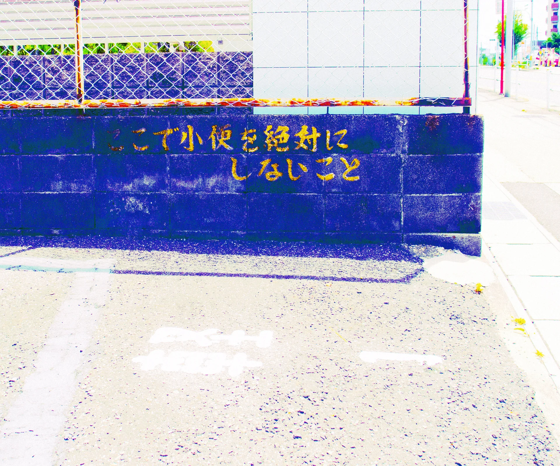 Yellow Japanese lettering marks a weathered blue boundary wall beneath wire mesh and pale panels.