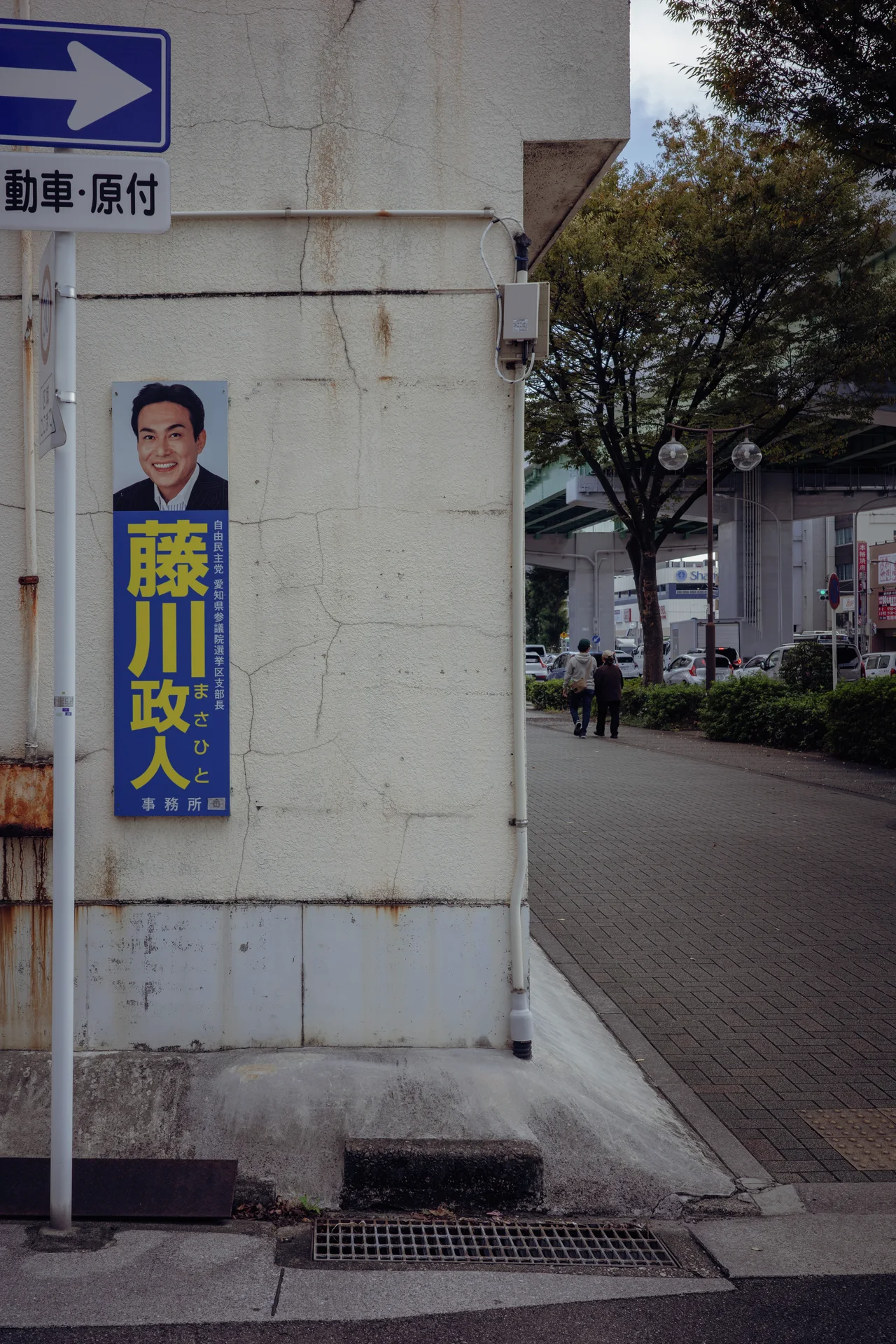 A portrait poster hangs on a pale corner wall beside a narrow tree-lined lane.