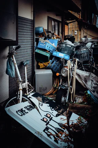 Bicycles and blue containers crowd a narrow passage beside a tilted sign and closed metal shutter.