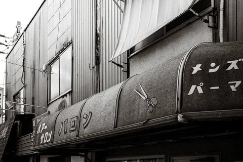 A curved barber shop awning displays scissors and Japanese lettering beneath corrugated walls in black and white.
