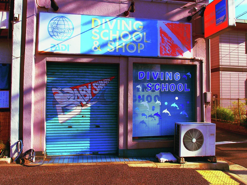 Blue diving-school signs and illustrated shutters mark a small shop beside an outdoor air conditioner.