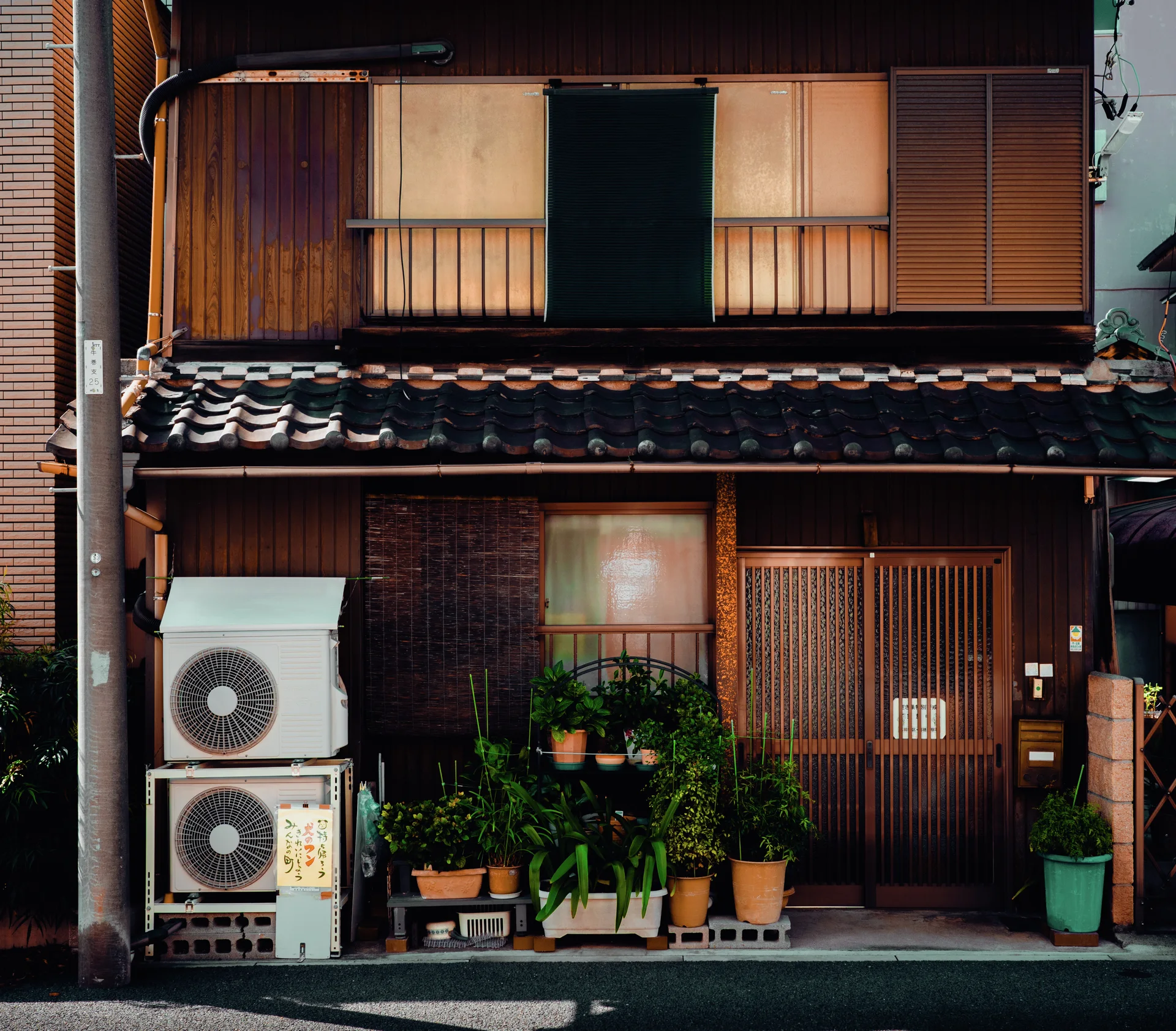 Potted greenery clusters beside two stacked air conditioners at a wooden house with a small tiled awning.