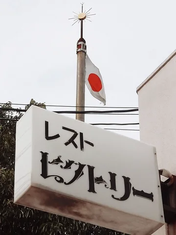 A pale projecting sign with black Japanese lettering hangs below a Japanese flag and slender spire.