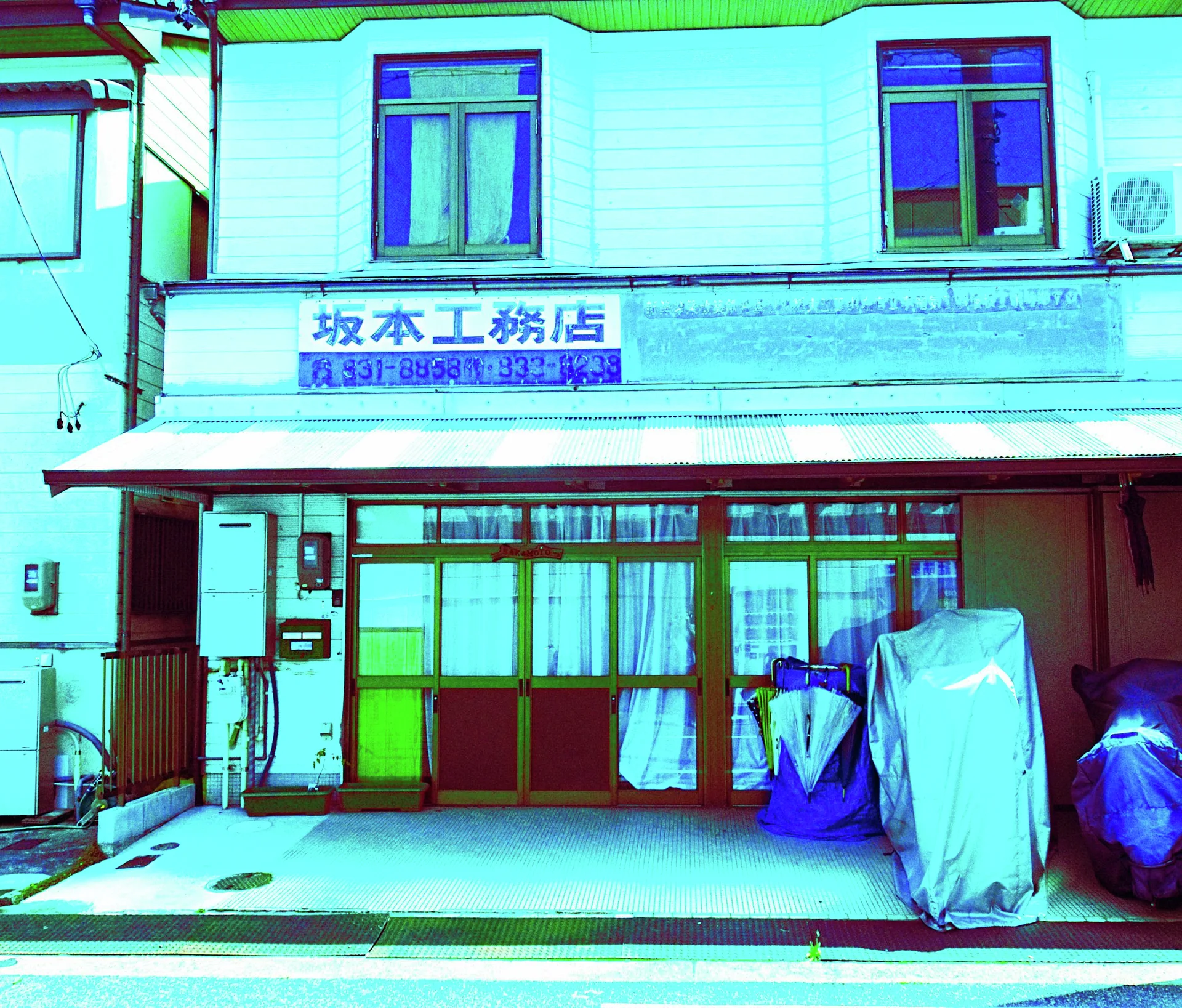 Covered objects stand outside a pale workshop with blue-framed windows and curtained sliding doors.