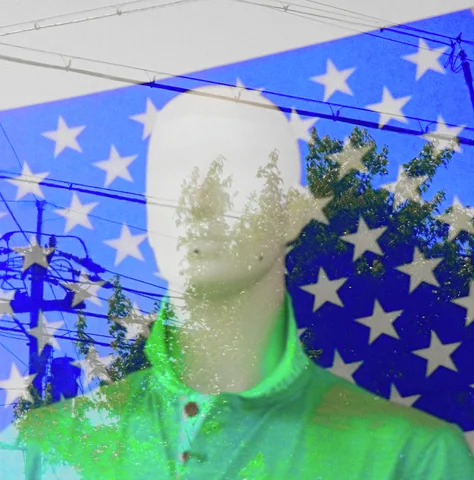 A pale mannequin in a green shirt stands against blue stars behind glass reflecting trees and wires.