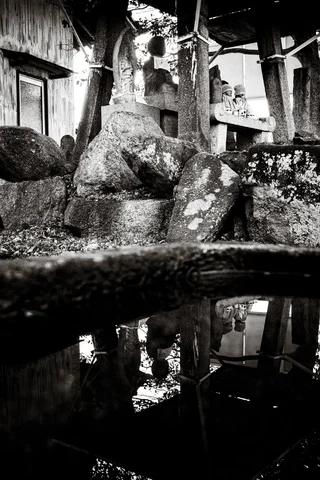 Rough stone forms and a tall pedestal reflect in a dark pool beside a building in black and white.