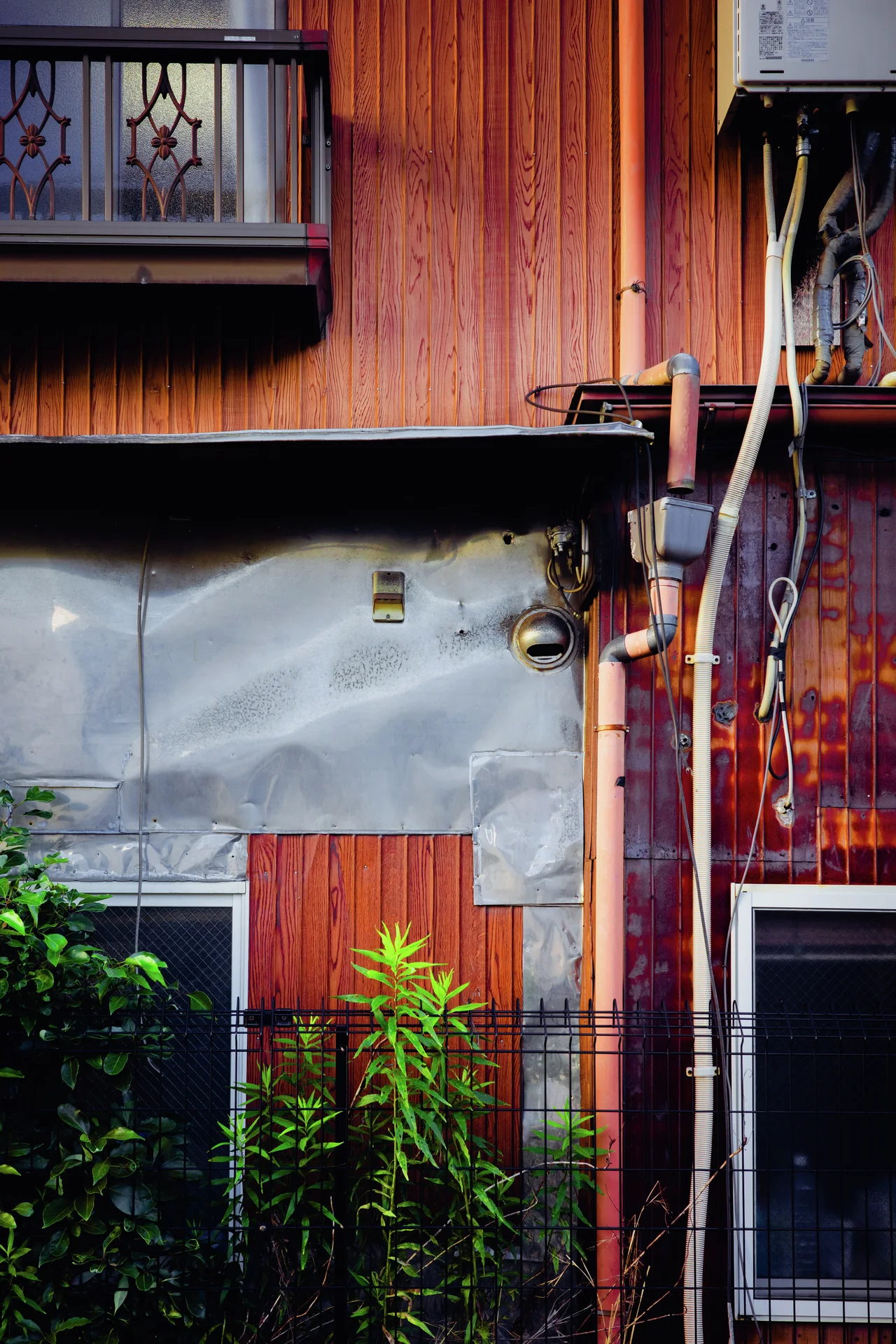 Exposed pipes and patchwork gray panels cover a rust-colored wall behind tall green plants.