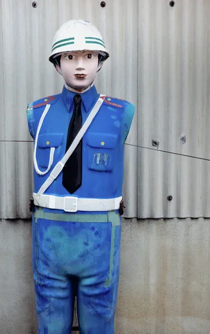 A weathered uniformed mannequin wears blue clothing and a white helmet against pale corrugated siding.