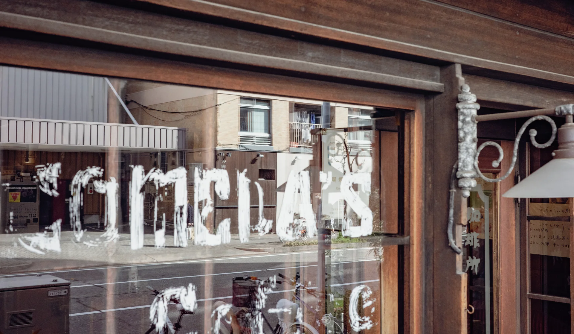 Distressed white lettering covers a wooden-framed shop window reflecting bicycles and buildings across the street.