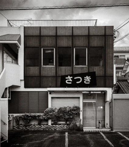 A dark two-story facade has twin upper windows and a pale sign above a shuttered entrance.