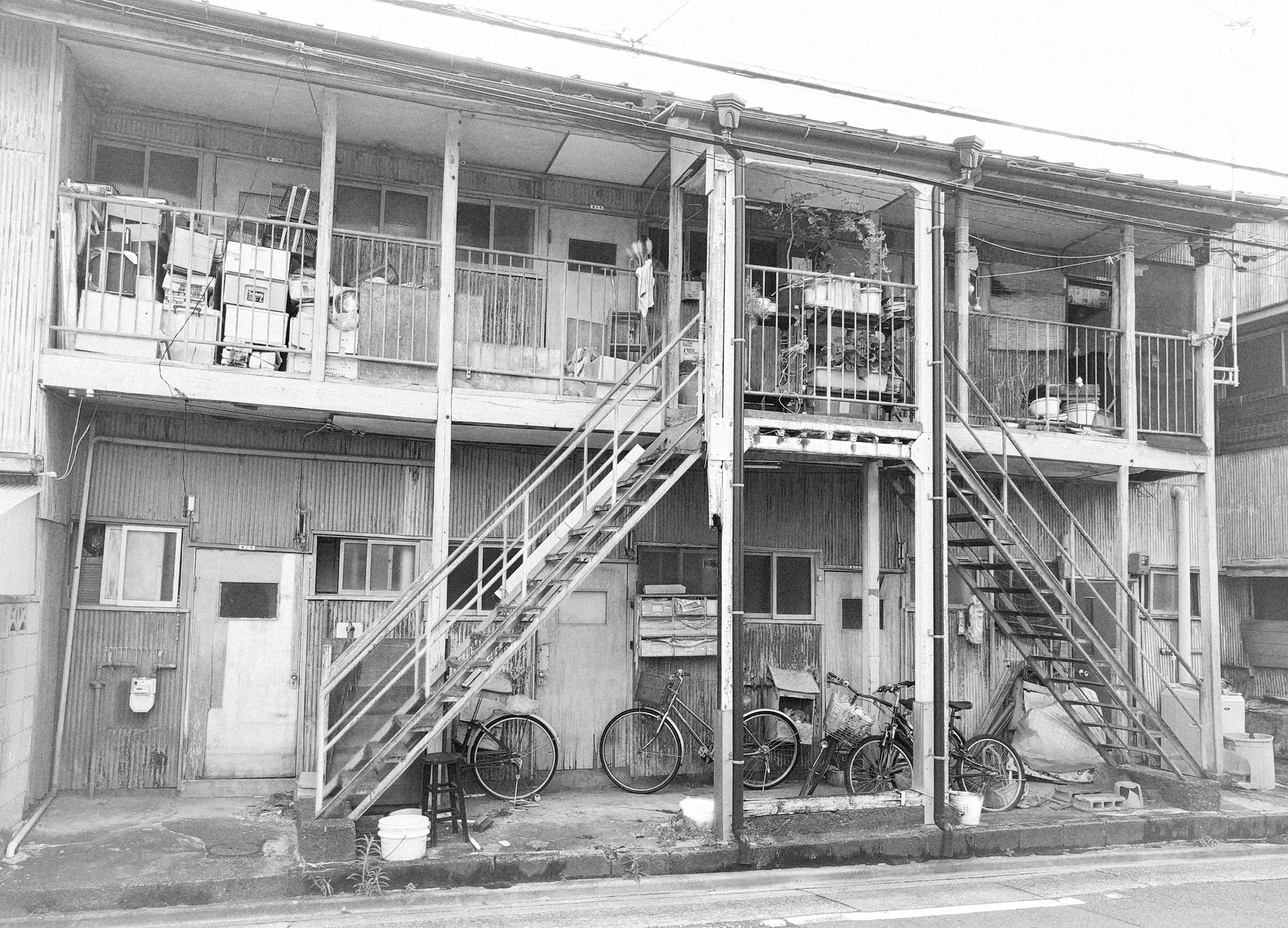 Two exterior staircases rise to a long apartment balcony above parked bicycles in black and white.