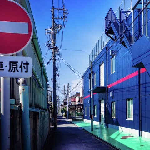 A narrow lane passes a vivid blue building with pink trim beside a no-entry sign.