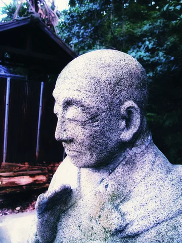 A weathered stone figure's bald head and calm face stand before a dark wooden structure and trees.