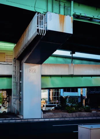 Turquoise elevated beams rest on pale concrete pillars beside a dark roadway and small industrial buildings.