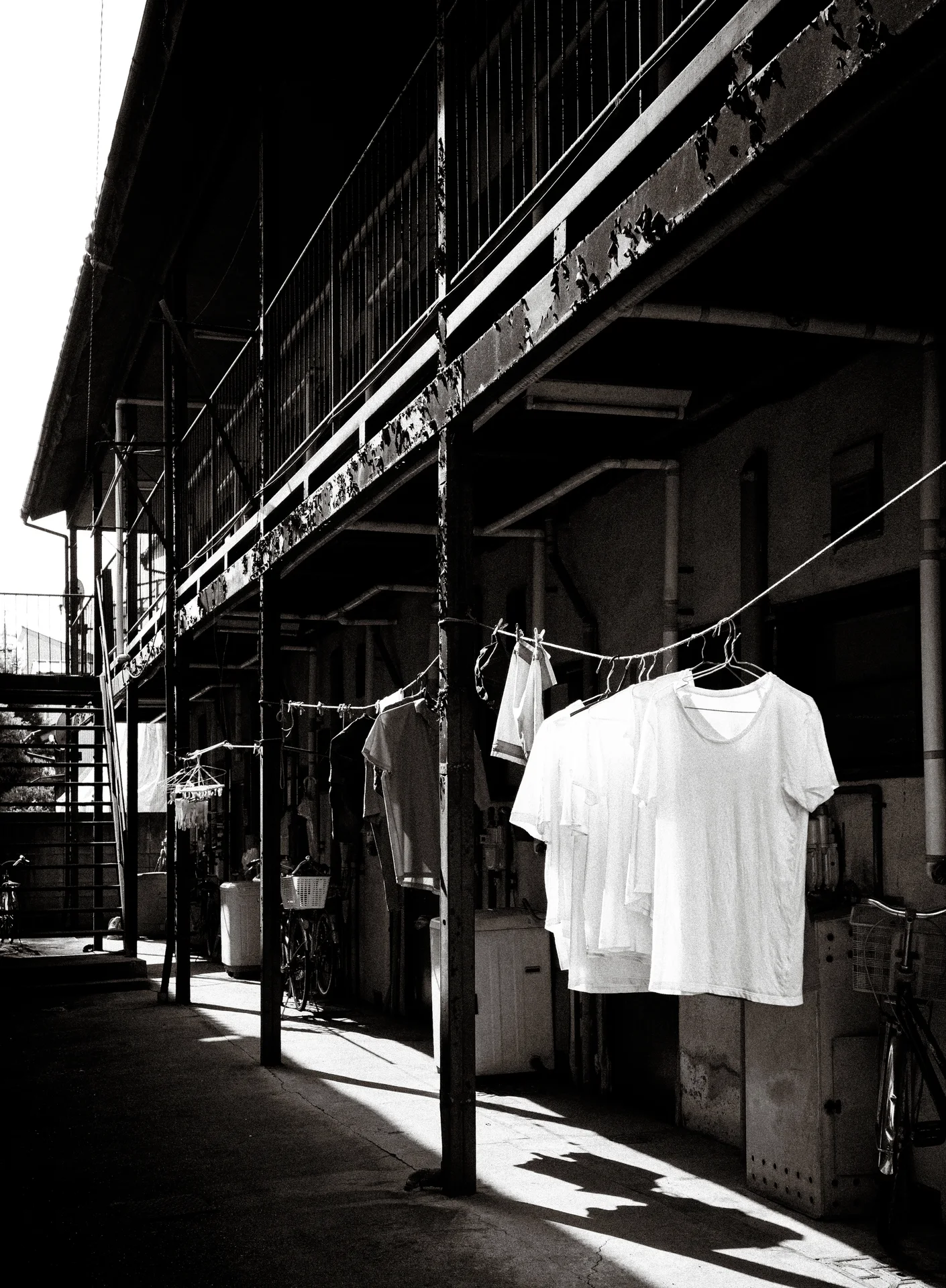 White shirts hang outside a long weathered building above strong sidewalk shadows in monochrome.