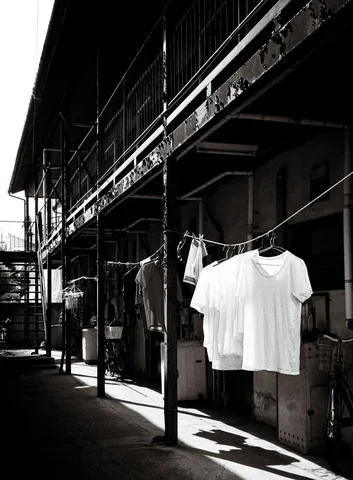 White shirts hang outside a long weathered building above strong sidewalk shadows in monochrome.
