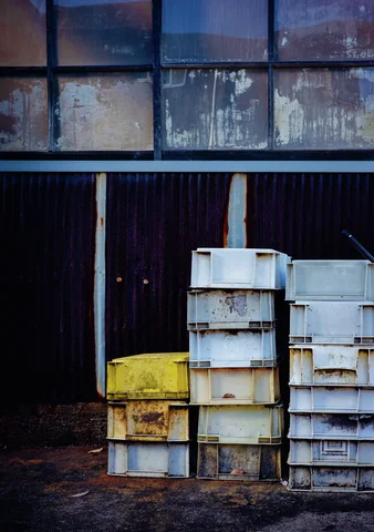 Tall stacks of weathered white and yellow crates stand against dark siding beneath stained upper windows.