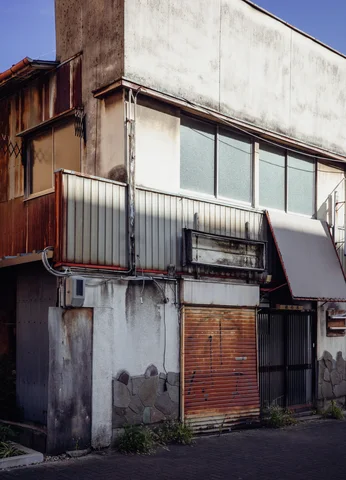 A weathered corner building has long upper windows, metal balcony panels and a closed rusty shutter.