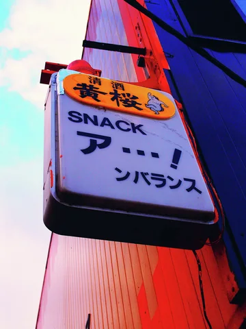 A pale projecting SNACK sign with Japanese lettering hangs beneath an orange panel beside red siding.