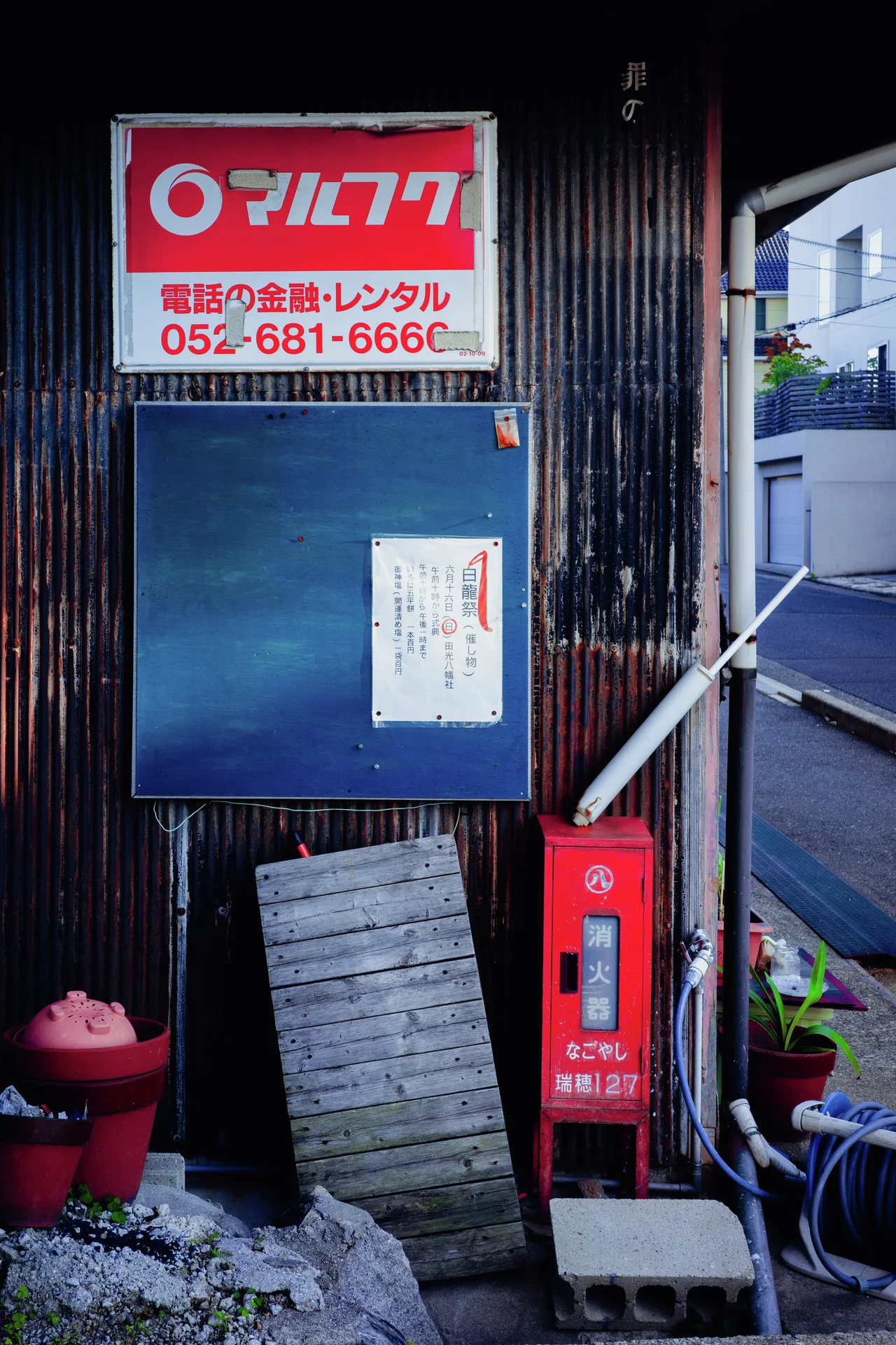 A red and white sign hangs above a blue noticeboard beside a red box and corrugated siding.