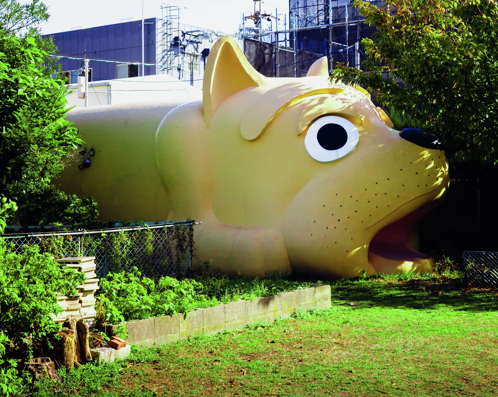 A large yellow animal-shaped structure with an open mouth stands amid shrubs behind a low wire fence.