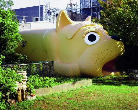 A large yellow animal-shaped structure with an open mouth stands amid shrubs behind a low wire fence.
