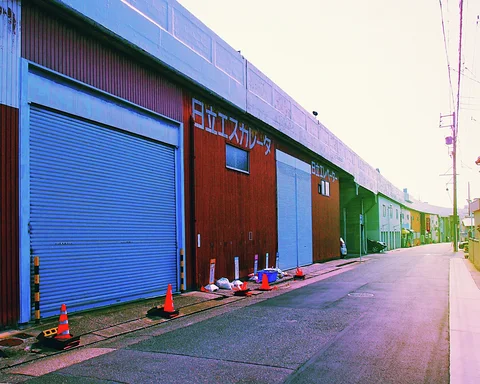 A long row of industrial buildings with blue shutters borders an empty paved lane and scattered cones.