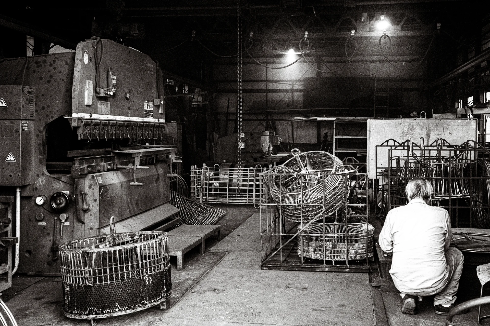 A crouching person faces stacked wire frames beside large machinery inside a workshop in black and white.