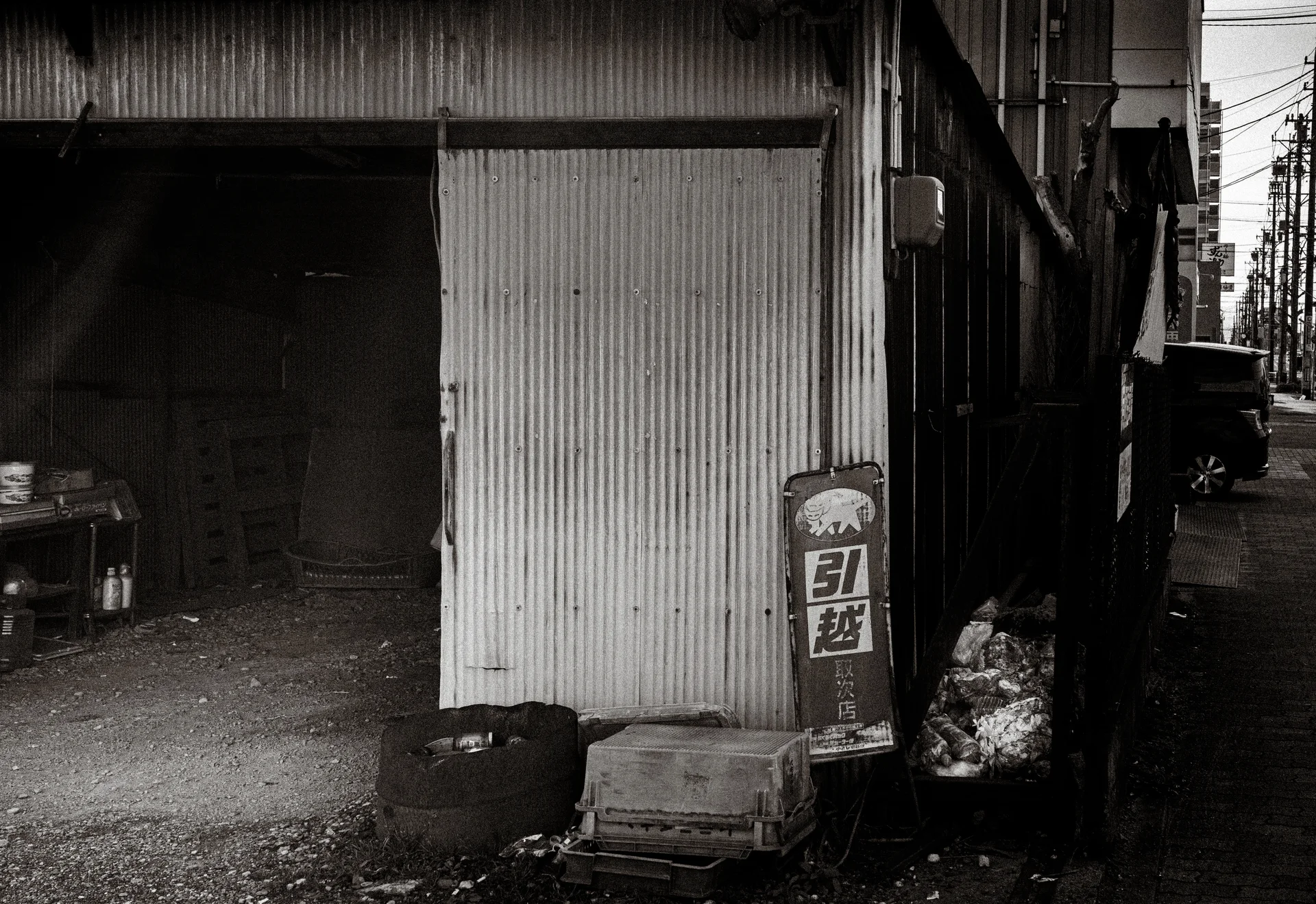 An open corrugated shed stands beside a narrow lane with stacked crates and sacks in black and white.
