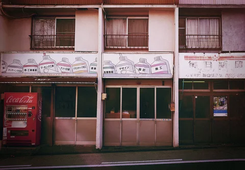 Faded illustrated shop signs span a two-story facade above sliding doors and a red vending machine.