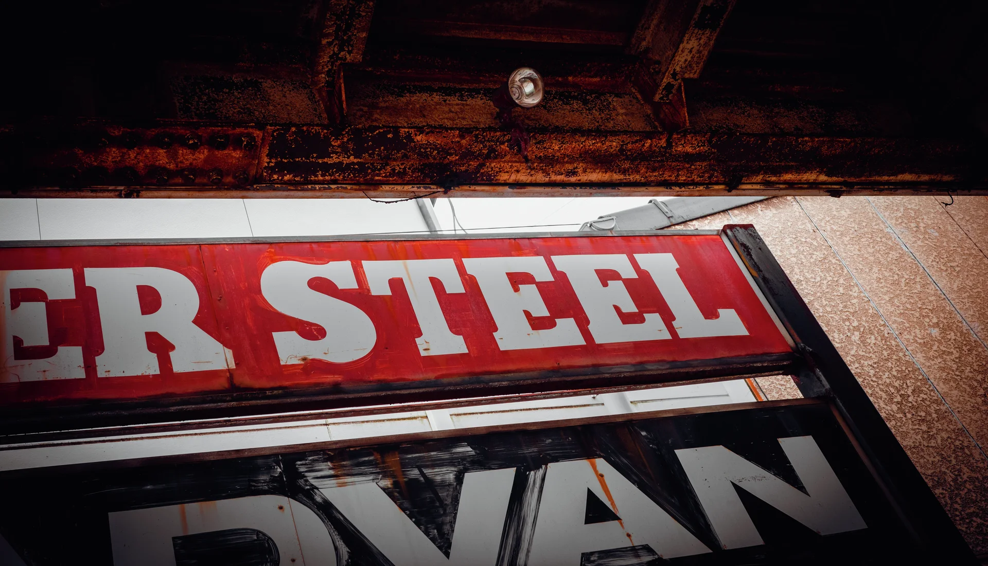 Large white Steel lettering fills a red sign beneath weathered timber and a small ceiling light.