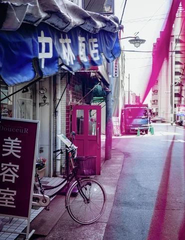 A bicycle rests beside shop signs and blue awning fabric in a narrow pink-toned passage.
