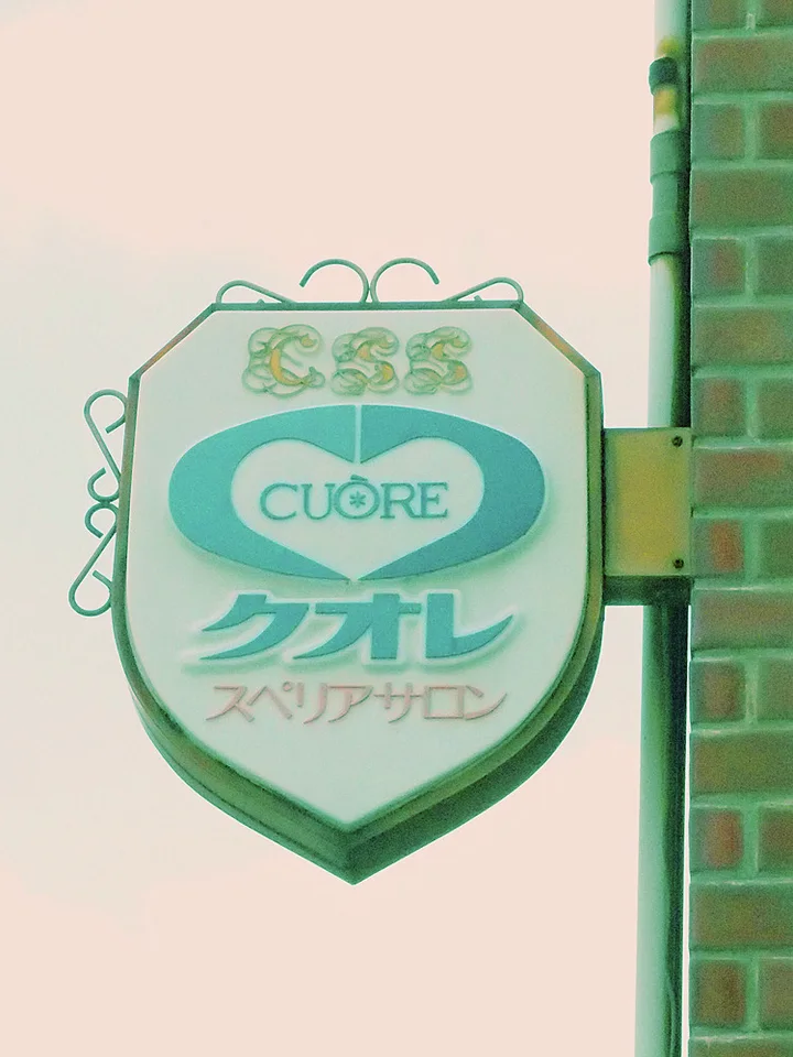 A pale shield-shaped CUORE hair salon sign hangs from decorative green metalwork beside brickwork.