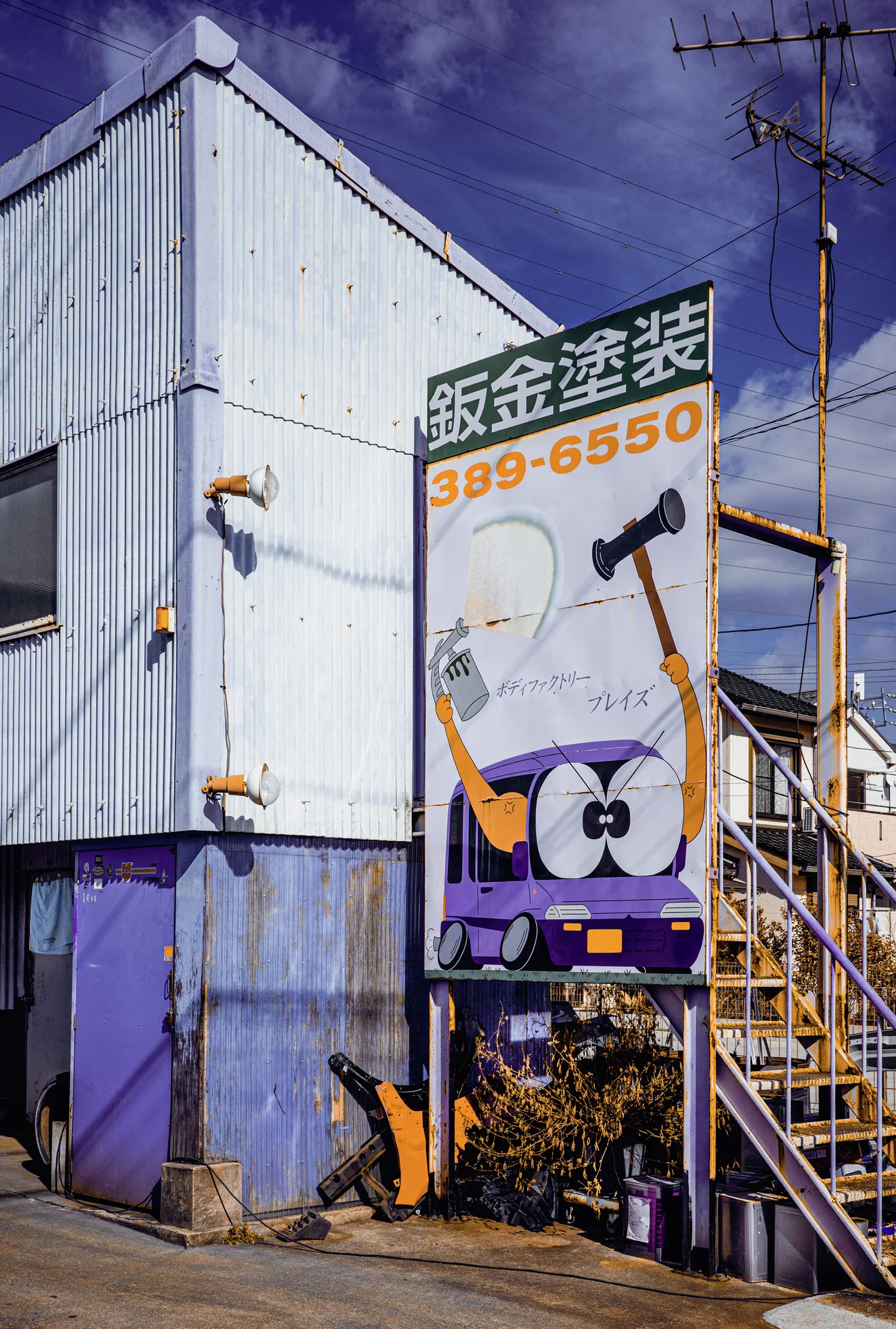 A tall sign with a cartoon car stands beside outdoor metal stairs on a corrugated building.