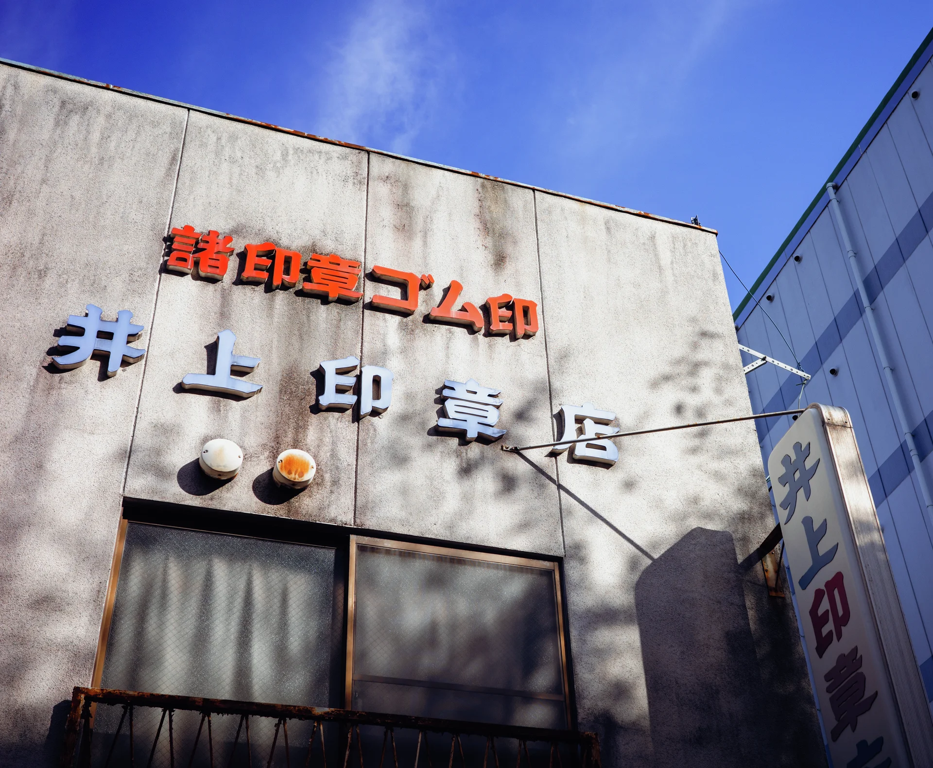 Raised orange and pale lettering decorates a stained building facade beneath blue sky and tree shadows.