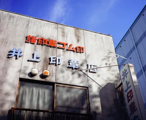 Raised orange and pale lettering decorates a stained building facade beneath blue sky and tree shadows.