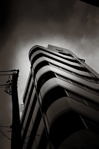 Curving balconies wrap around a tall building seen from below beneath dark clouds in black and white.