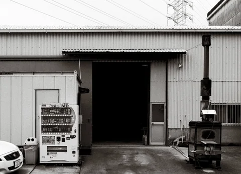 A vending machine stands beside an open warehouse bay and a tall metal chimney in black and white.