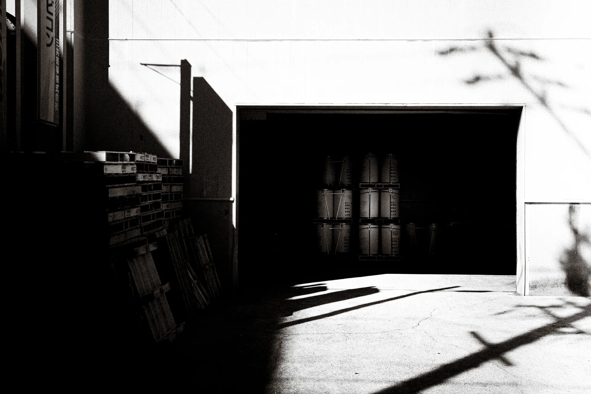 A sunlit industrial entrance reveals stacked cylinders inside darkness, crossed by strong diagonal shadows.