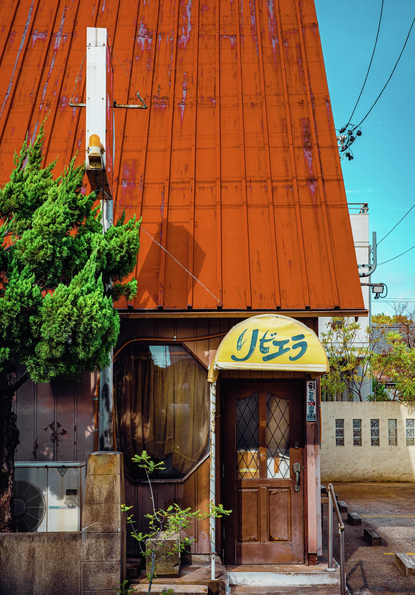 A steep orange metal facade towers above a narrow wooden entrance and a small yellow sign.